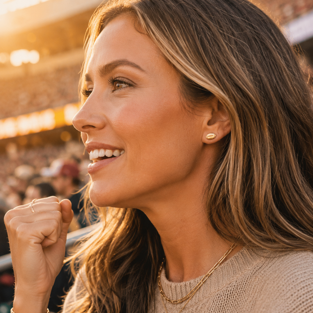A woman in a stadium wearing a small gold stud earring and layered gold necklaces, captured in warm golden-hour lighting.