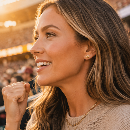 A woman in a stadium wearing a small gold stud earring and layered gold necklaces, captured in warm golden-hour lighting.