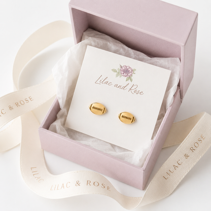 A pair of small gold football-shaped stud earrings displayed on a white card inside a pale pink gift box, with a cream ribbon printed with the brand name Lilac and Rose.