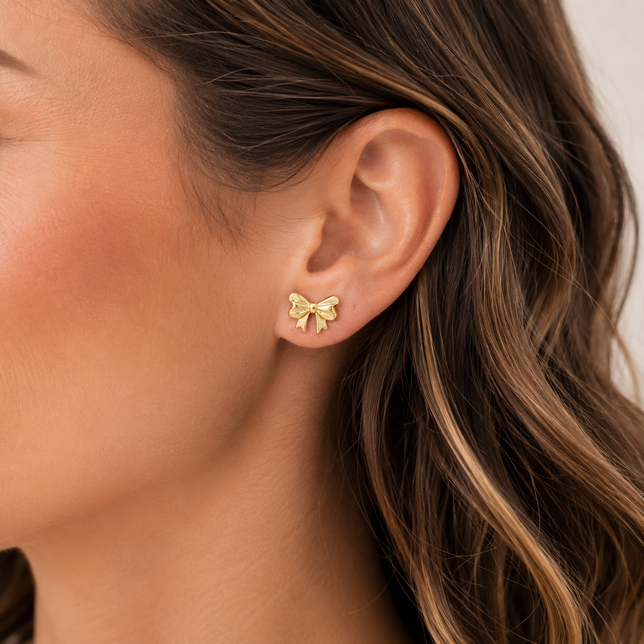 A close-up of a woman's ear wearing a small gold bow stud earring, with her brown hair visible against a light neutral background.
