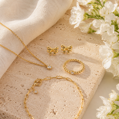 A gold jewelry set arranged on a textured stone slab with white fabric and white flowers. The set includes a delicate chain necklace with a small clear stone pendant, a bracelet, a ring, and two bow-shaped stud earrings.