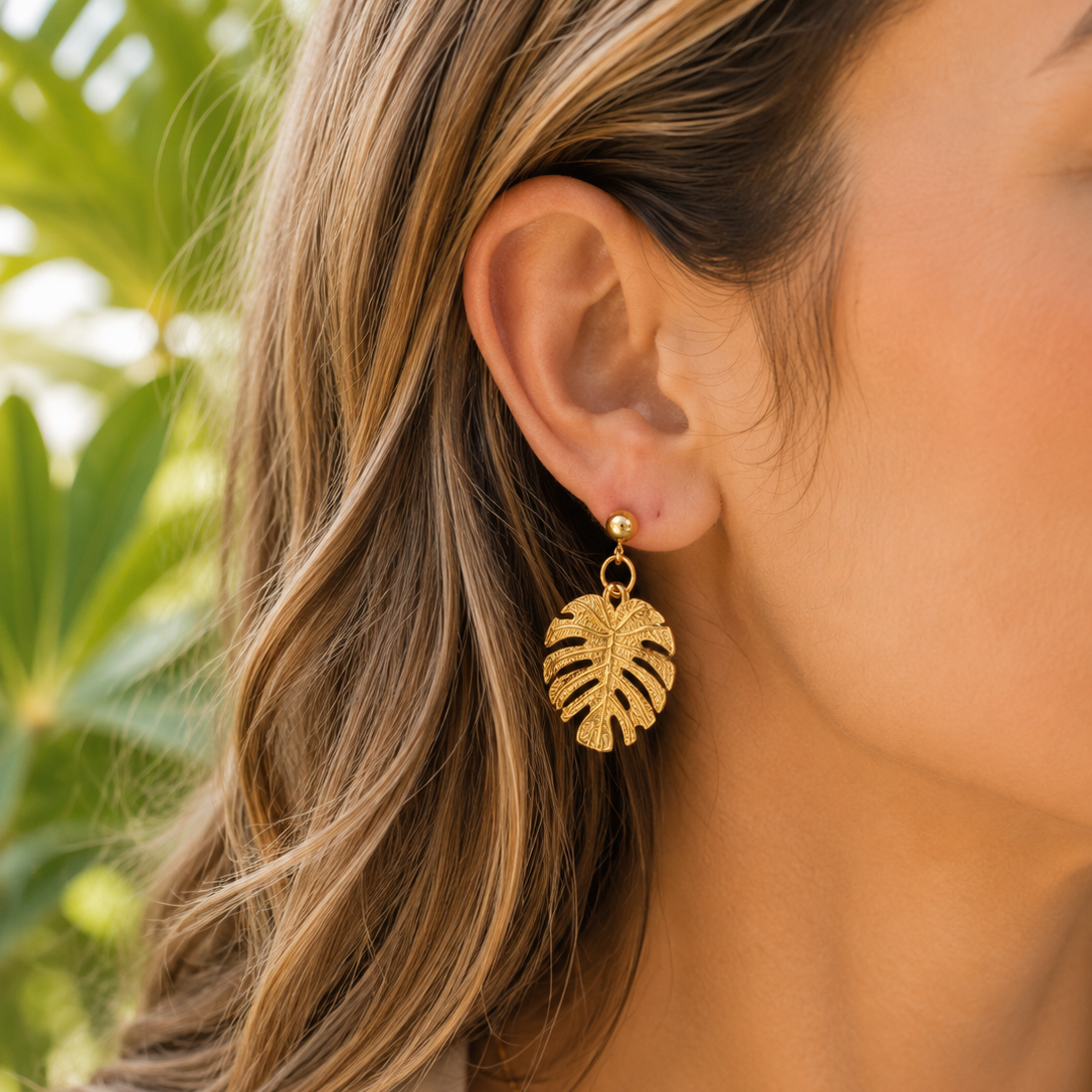 A close-up of a woman wearing a gold leaf drop earring, with her ear, hair, and neck visible against a soft-focus green outdoor background.