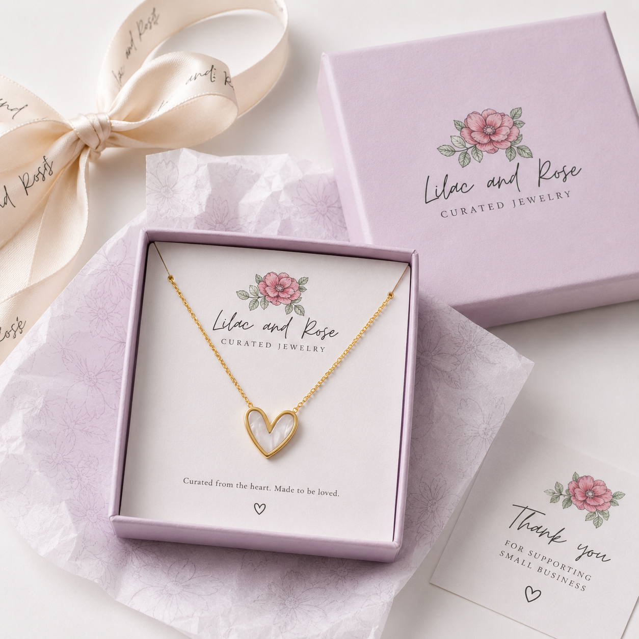 A gold heart-shaped necklace with a hollow center is displayed in a lavender jewelry gift box. The box is branded 'Lilac and Rose Curated Jewelry' with a pink rose illustration.