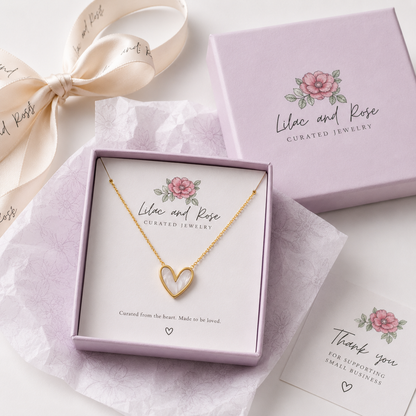 A gold heart-shaped necklace with a hollow center is displayed in a lavender jewelry gift box. The box is branded 'Lilac and Rose Curated Jewelry' with a pink rose illustration.