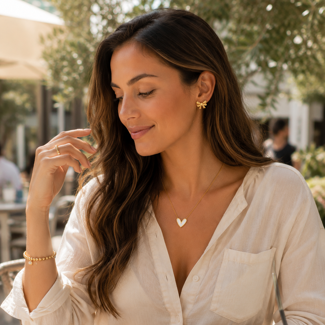 A woman wearing a delicate gold heart pendant necklace, a gold heart stud earring, and a gold bracelet, posing in a light-colored linen blouse in an outdoor patio setting.