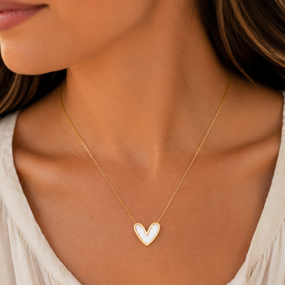 A close-up of a woman wearing a delicate gold chain necklace with a small white heart-shaped pendant.