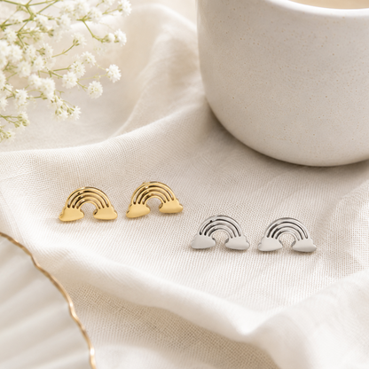 Three small rainbow-shaped stud earrings arranged on cream fabric, with two gold-toned pairs on the left and one silver-toned pair on the right. A white ceramic cup and white baby's breath flowers are softly blurred in the background.