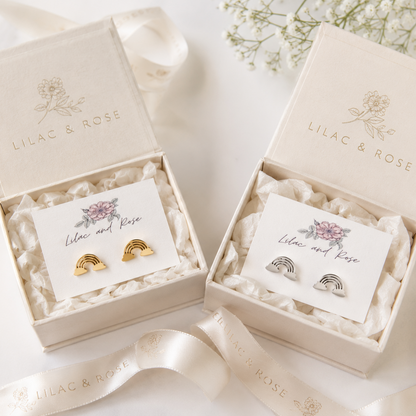 Two open cream-colored Lilac & Rose gift boxes on a white surface, each holding a small jewelry card with rainbow-shaped earrings. One pair is gold and the other is silver, surrounded by white satin ribbon and delicate white flowers.