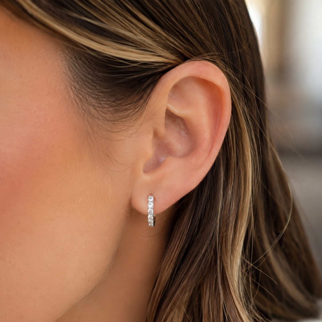 A close-up of a woman's ear wearing a small silver huggie hoop earring set with clear stones.