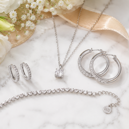 A jewelry set arranged on a white marble surface with white flowers and a satin ribbon. The set includes a silver-toned necklace with a heart-shaped clear stone pendant, matching hoop earrings, a tennis bracelet, and a smaller hoop earring.