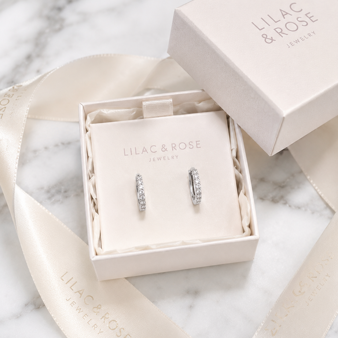 A pair of silver hoop earrings with clear stones, presented in an open cream-colored jewelry box on a cream display card. The box lid and a matching cream satin ribbon with 'LILAC & ROSE JEWELRY' branding are arranged on a white marble surface.