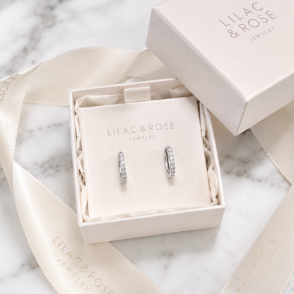 A pair of silver hoop earrings with clear stones, presented in an open cream-colored jewelry box on a cream display card. The box lid and a matching cream satin ribbon with 'LILAC & ROSE JEWELRY' branding are arranged on a white marble surface.