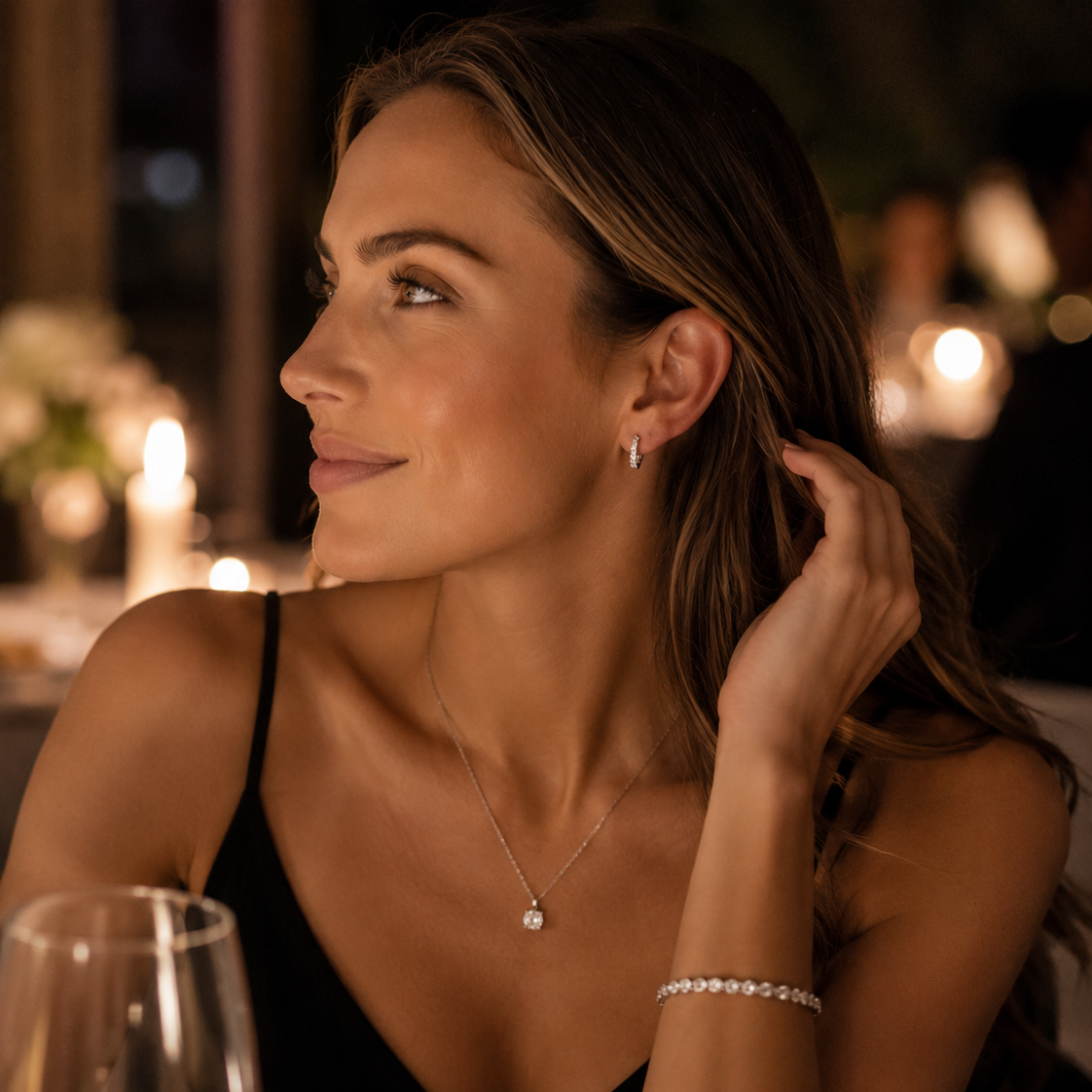 A woman in a black dress wearing a delicate silver necklace with a small clear stone pendant and a matching silver bracelet, photographed in a warmly lit restaurant setting.