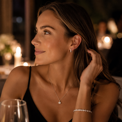 A woman in a black dress wearing a delicate silver necklace with a small clear stone pendant and a matching silver bracelet, photographed in a warmly lit restaurant setting.