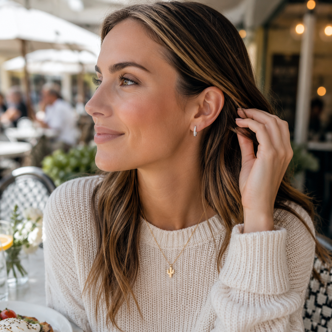 A woman with long brown hair sits at an outdoor cafe table wearing a cream knit sweater and a delicate gold necklace with a small letter pendant. A plate of avocado toast is visible on the table in the foreground.