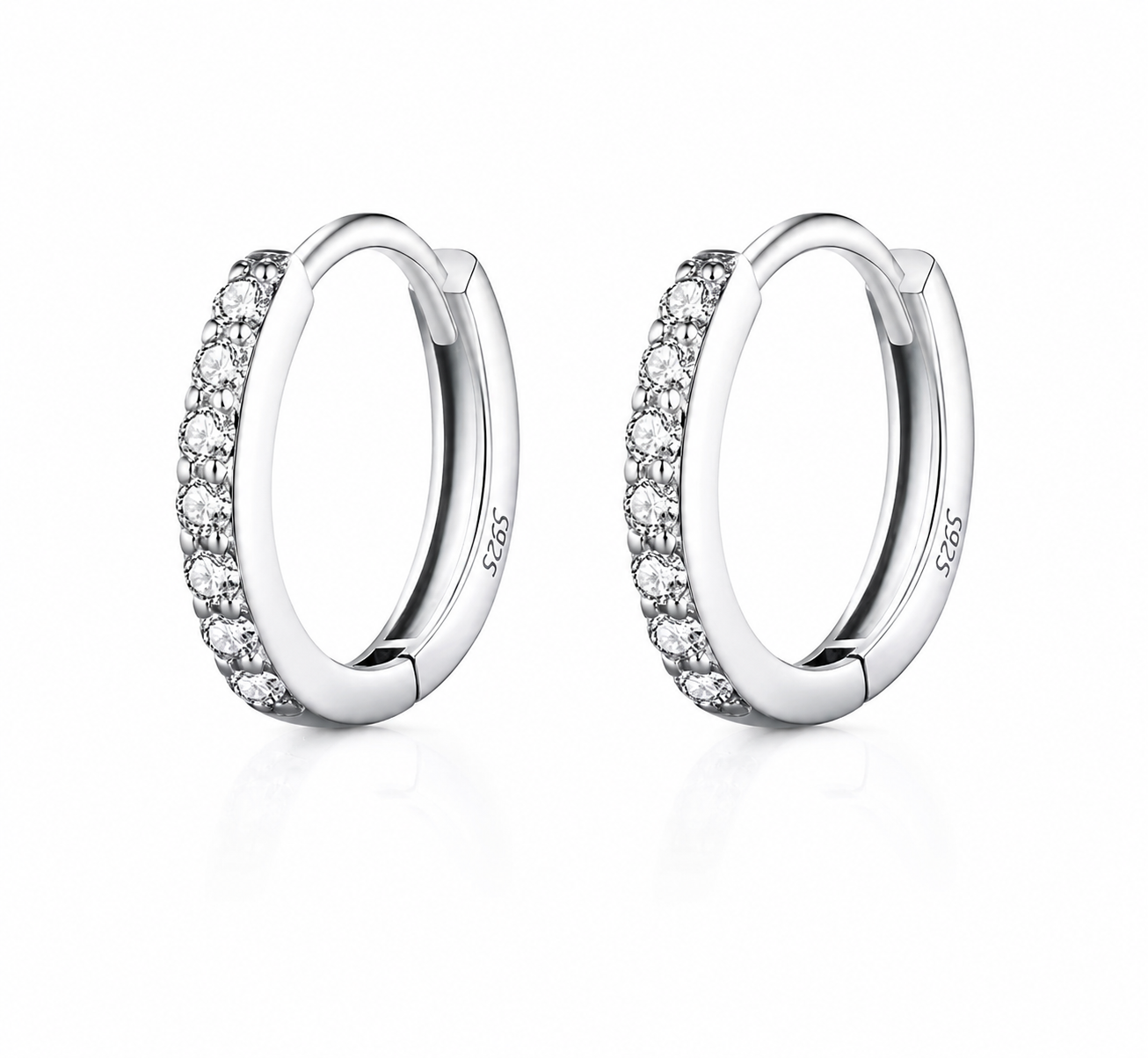 A pair of silver huggie hoop earrings with a row of clear pavé-set round stones on the front, shown on a clean white background with a soft reflection.