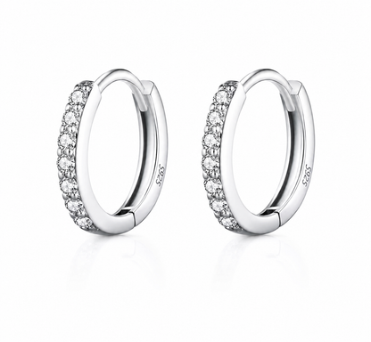 A pair of silver huggie hoop earrings with a row of clear pavé-set round stones on the front, shown on a clean white background with a soft reflection.