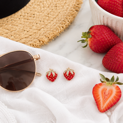 Red strawberry-shaped stud earrings with white pearl-like details and gold-tone tops, styled on a white fabric surface with sunglasses, a straw hat, strawberries, and a bowl of fresh strawberries.