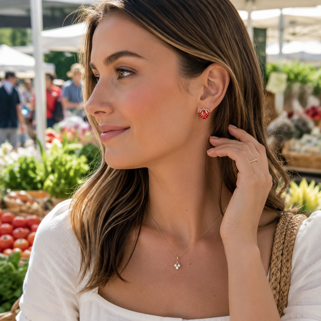 A woman wearing a delicate gold necklace with a small cross pendant and matching red heart stud earrings, photographed at a sunny outdoor farmers market.