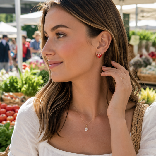 A woman wearing a delicate gold necklace with a small cross pendant and matching red heart stud earrings, photographed at a sunny outdoor farmers market.
